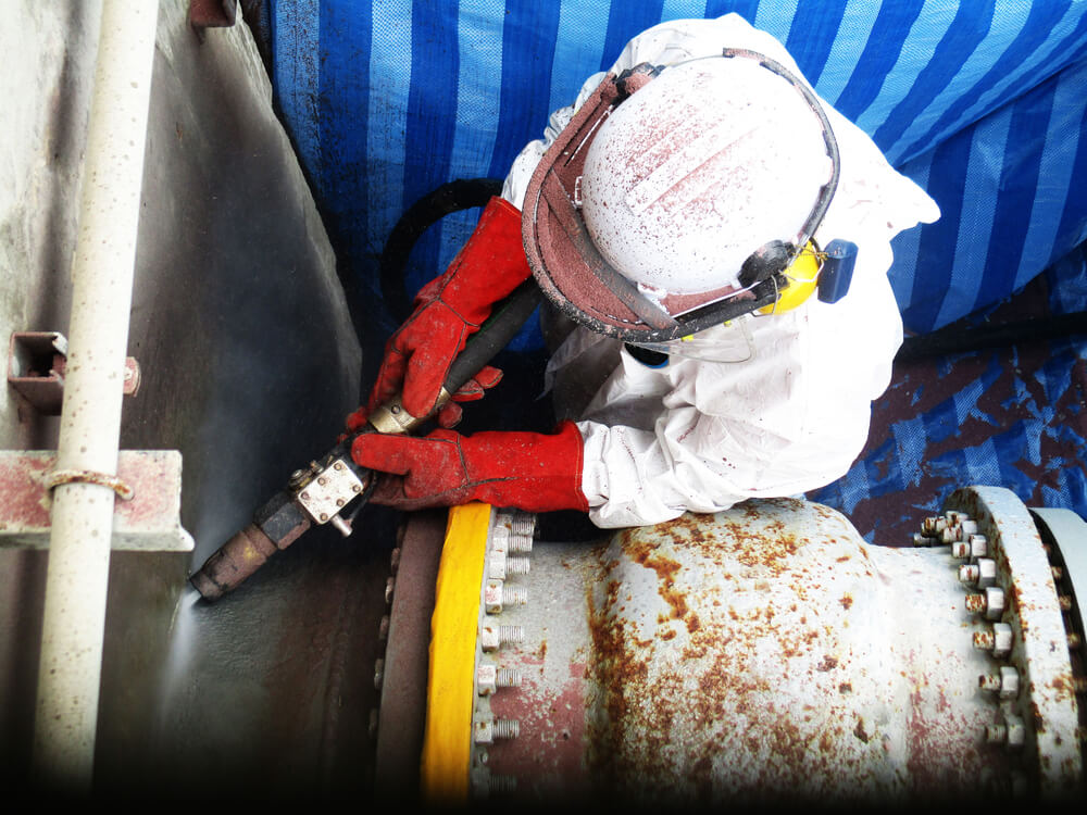 A technician fully equipped with protective gear, operating dustless blasting equipment to clean and restore a surface.